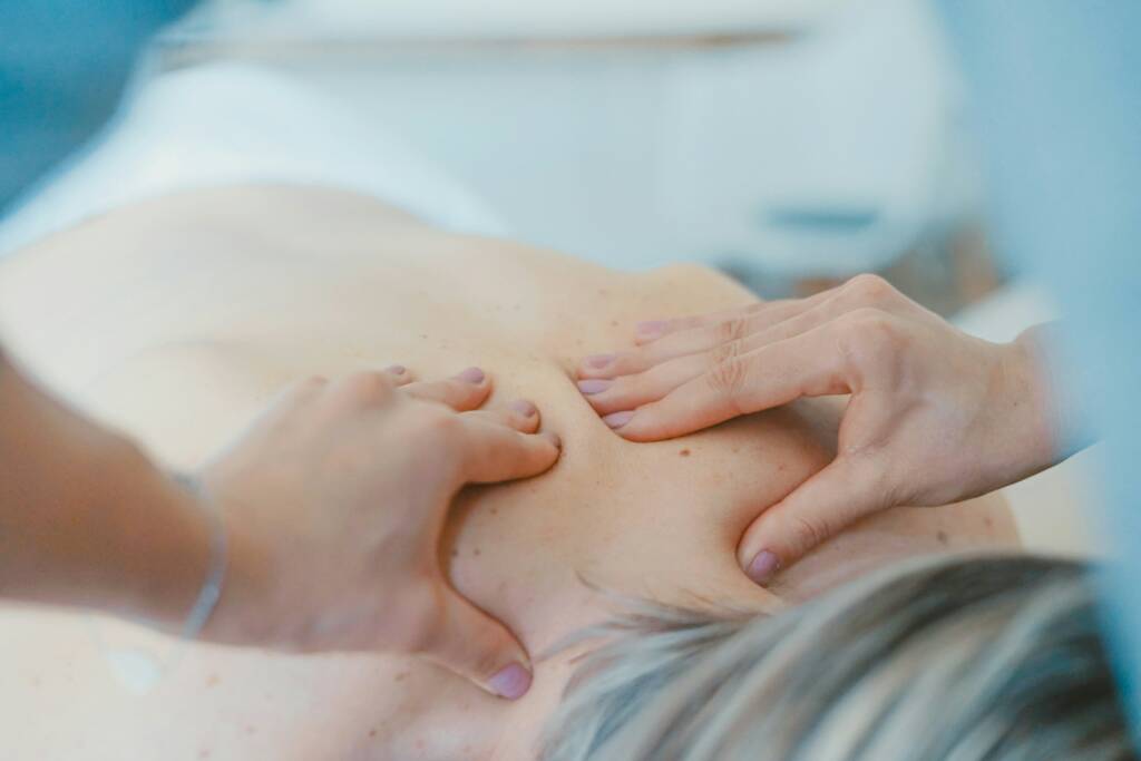 L'image montre une séance de massage Deep Tissue. On y voit une personne allongée sur le ventre, recevant un massage sur la partie supérieure de son dos. Le masseur applique une pression profonde avec ses mains sur les muscles du dos du patient, en utilisant ses doigts et ses paumes pour cibler des zones spécifiques. Le but de ce type de massage est de relâcher les tensions musculaires et les nœuds en pénétrant profondément dans les couches musculaires. La peau de la personne massée est légèrement rouge, signe de la pression appliquée. L'ambiance semble calme et relaxante, avec une attention particulière aux besoins du patient.